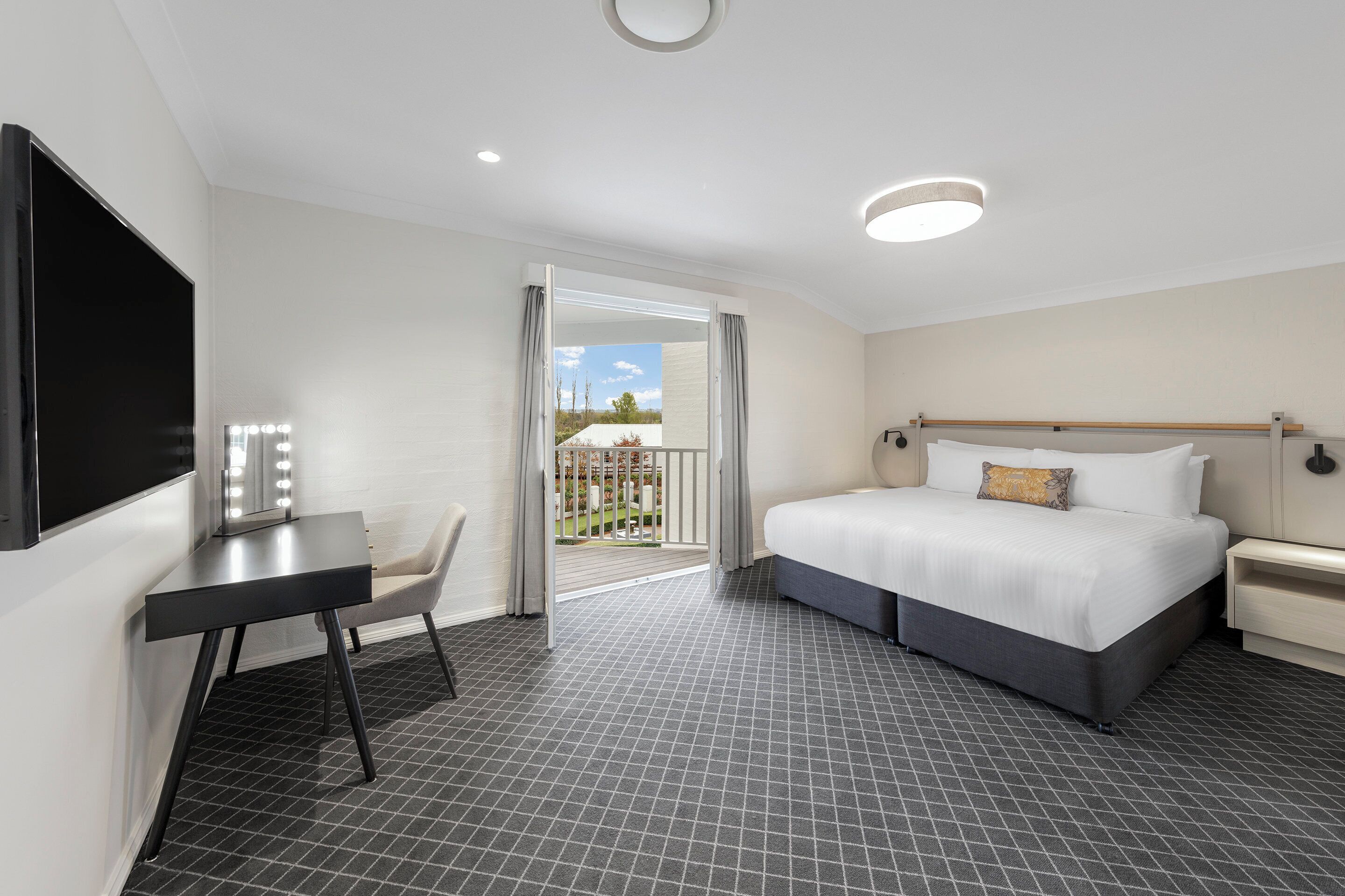 A hotel room with a bed, a desk with a chair, a TV, and a balcony with a view.