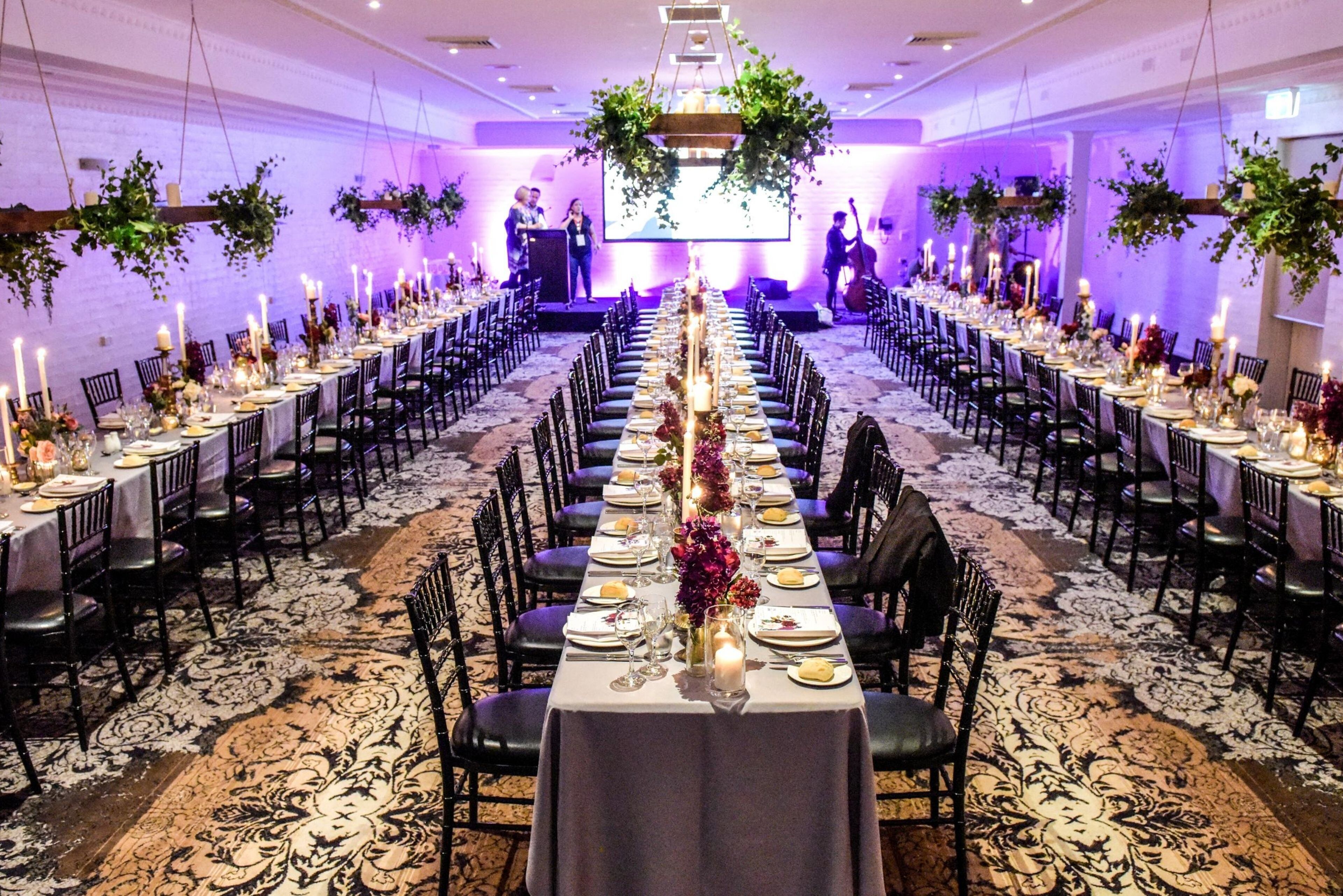 Un salón de banquetes con largas mesas dispuestas para un evento formal, que cuenta con un escenario central y un fondo decorado con follaje.