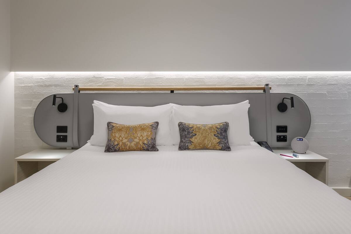 Een netjes opgemaakt bed met twee kussens, een nachtkastje met een lamp en een kleine speaker.