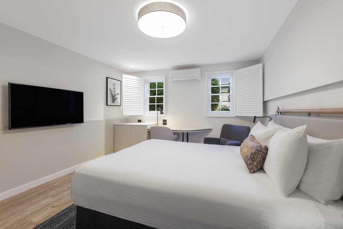 Een moderne hotelkamer met een groot bed, een flatscreen-televisie, een bureau en twee ramen met zonwering.