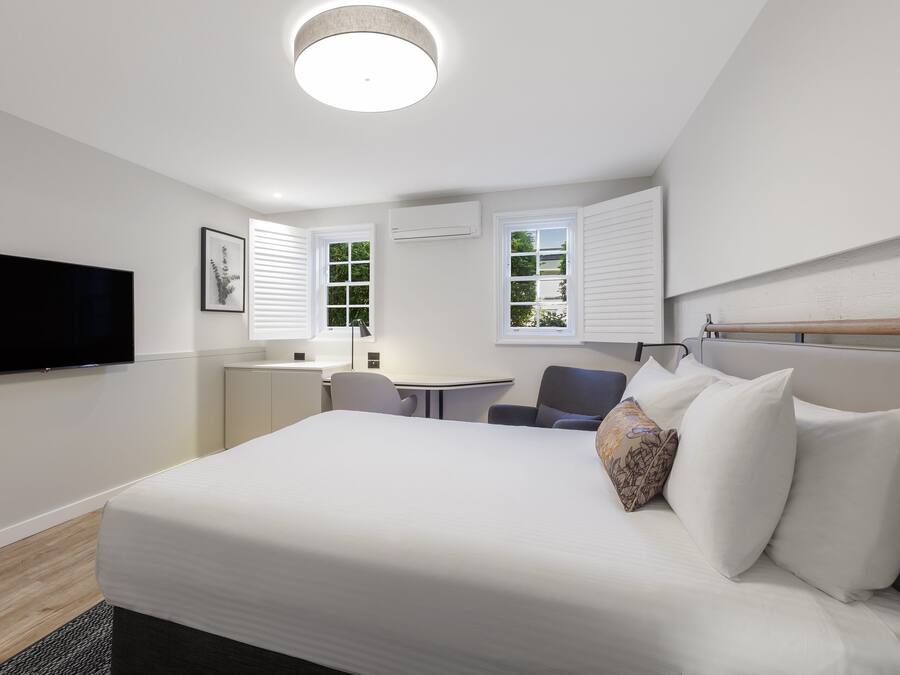 A modern hotel room with a large bed, a flat-screen TV, a desk, and two windows with blinds.