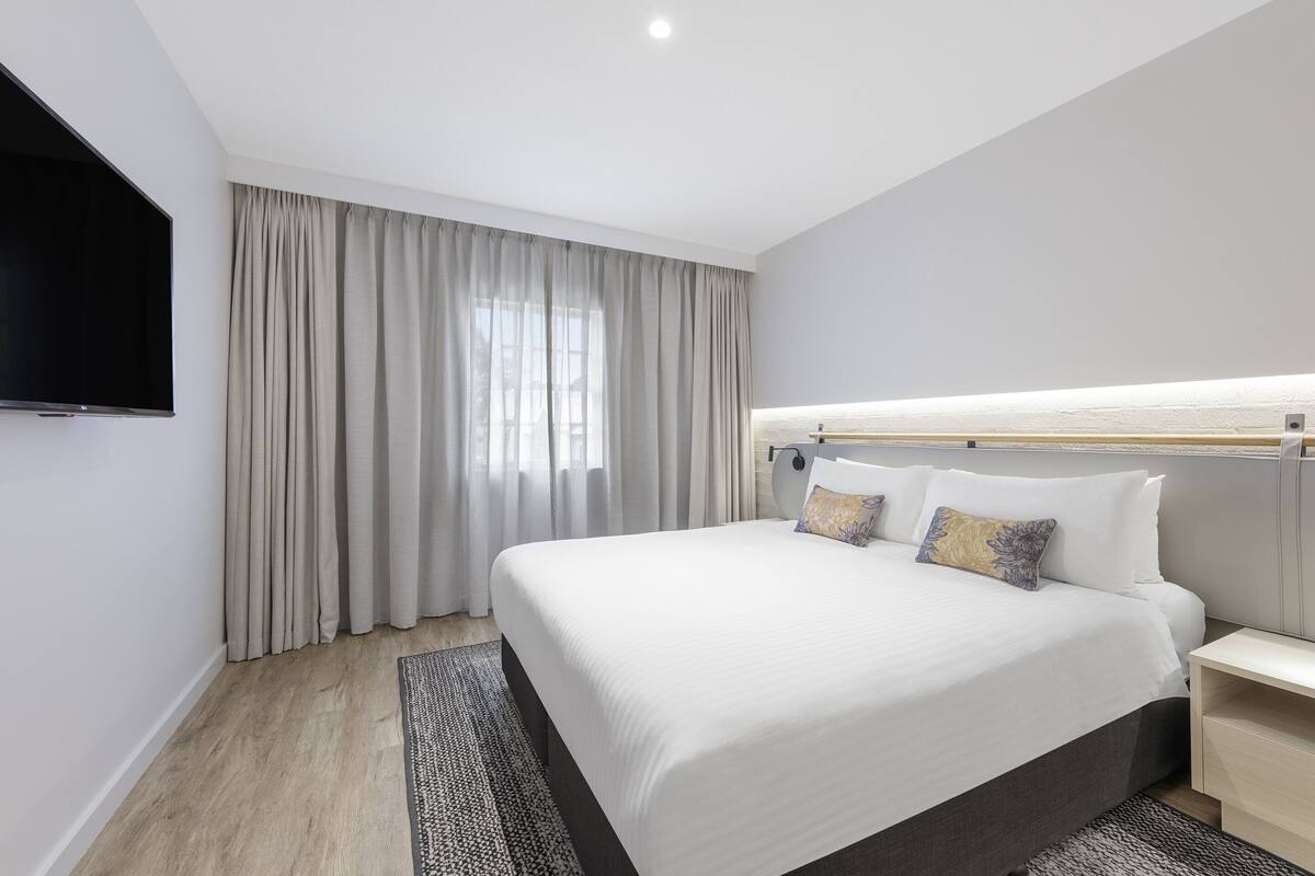 Een moderne hotelkamer met een groot bed, een flatscreen televisie aan de muur en een groot raam met gordijnen.