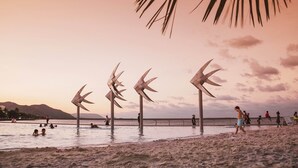 A beach with people swimming and walking, palm trees, and wind sculptures. - Hides Hotel Cairns (Cairns)