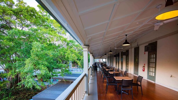 A covered outdoor seating area with wooden tables and chairs, a railing, and a view of greenery and trees. - Hides Hotel Cairns (Cairns)