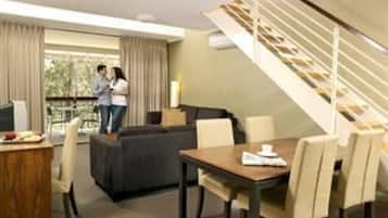 A hotel room with a sofa, dining table, chairs, a staircase, and a view of the outdoors.