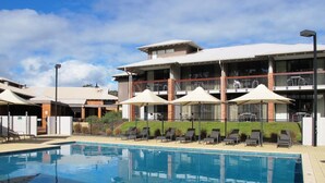 A swimming pool with lounge chairs and umbrellas, a building with a flat roof and large windows, and a clear sky. - Margarets in Town Apartments (Margaret River)