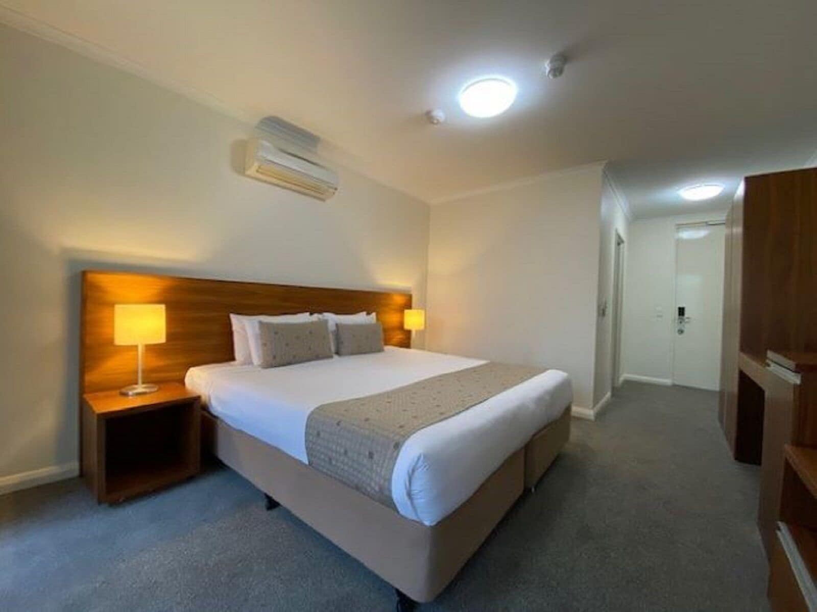 Ein Hotelzimmer mit einem großen Bett, Nachttischlampen, einer Deckenleuchte und einer Tür zu einem weiteren Raum.