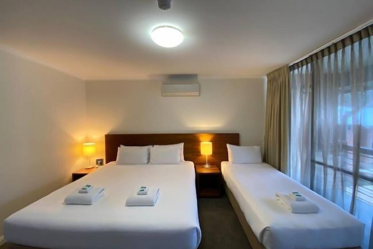 A hotel room with two single beds, each with a bedside table and lamp, a ceiling light, and a window with curtains.