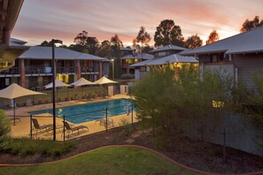 A swimming pool with lounge chairs and umbrellas, surrounded by buildings with balconies and a sunset sky. - Margarets in Town Apartments (Margaret River)