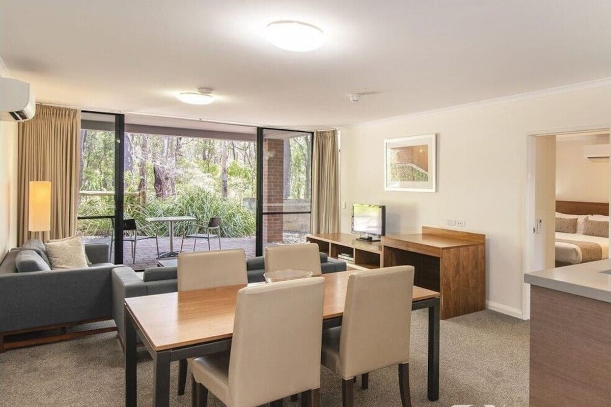 A modern hotel room with a dining area, a kitchen, and a balcony with a view of the outdoors.