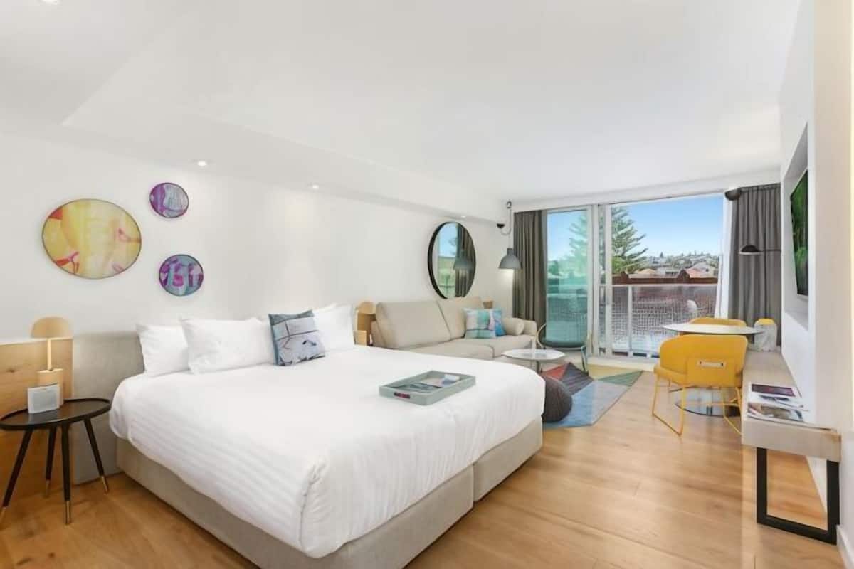 A modern hotel room with a large bed, a sofa, a small table, and a chair. There is a balcony with a view of the outside.