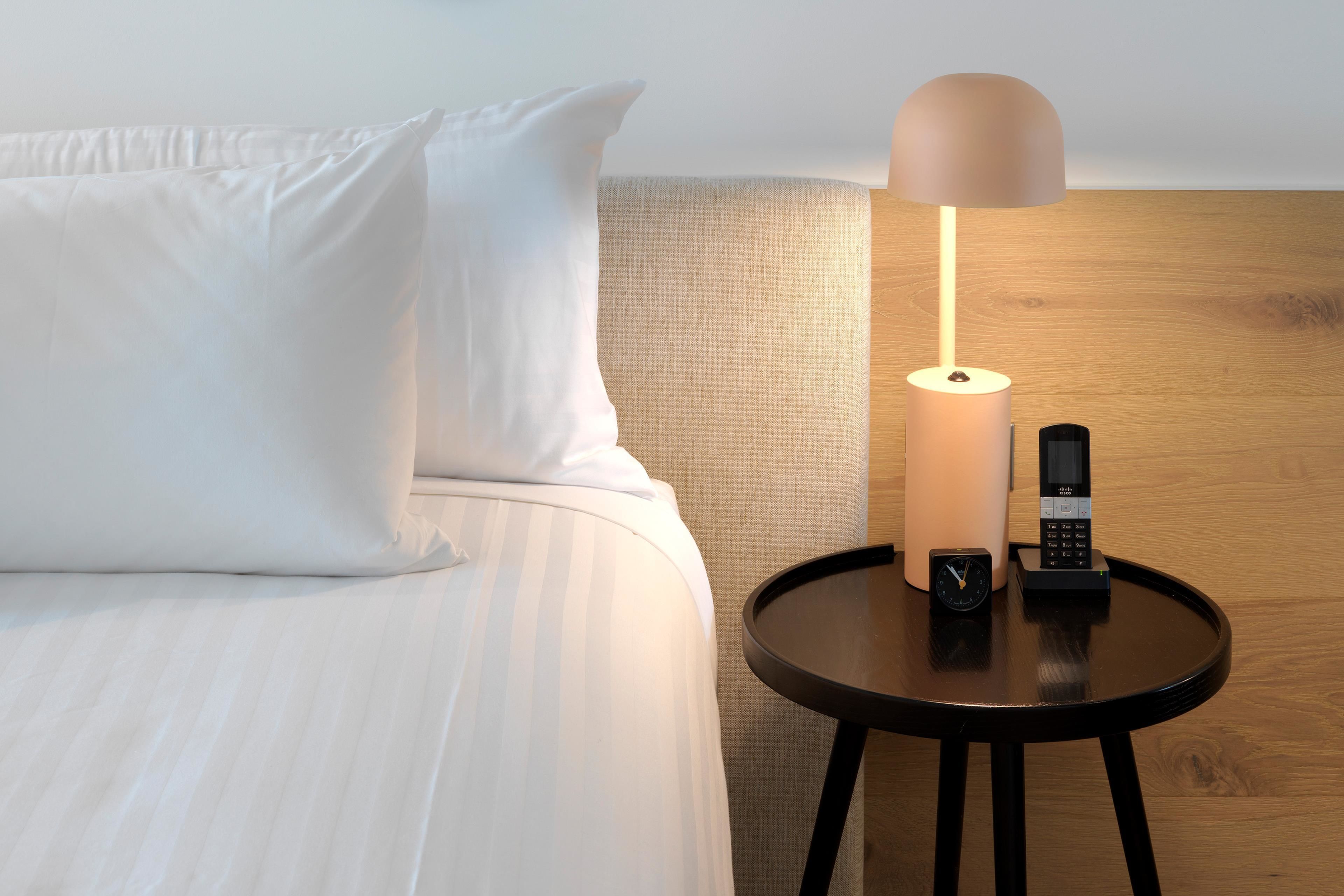 A neatly made bed with white linens, a round wooden nightstand with a lamp, and a telephone.