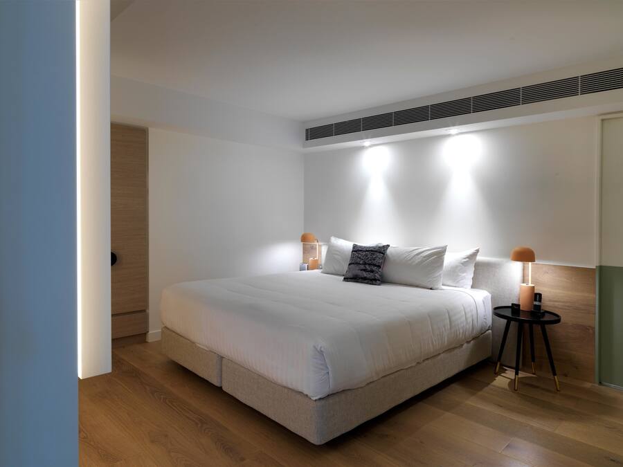 Une chambre moderne avec un grand lit, deux tables de chevet et une bouche d’aération au plafond.