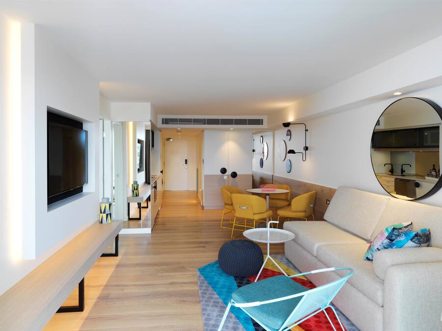 Un soggiorno moderno con una TV a schermo piatto, un divano, poltrone gialle, uno specchio rotondo e un piccolo tavolo da pranzo.