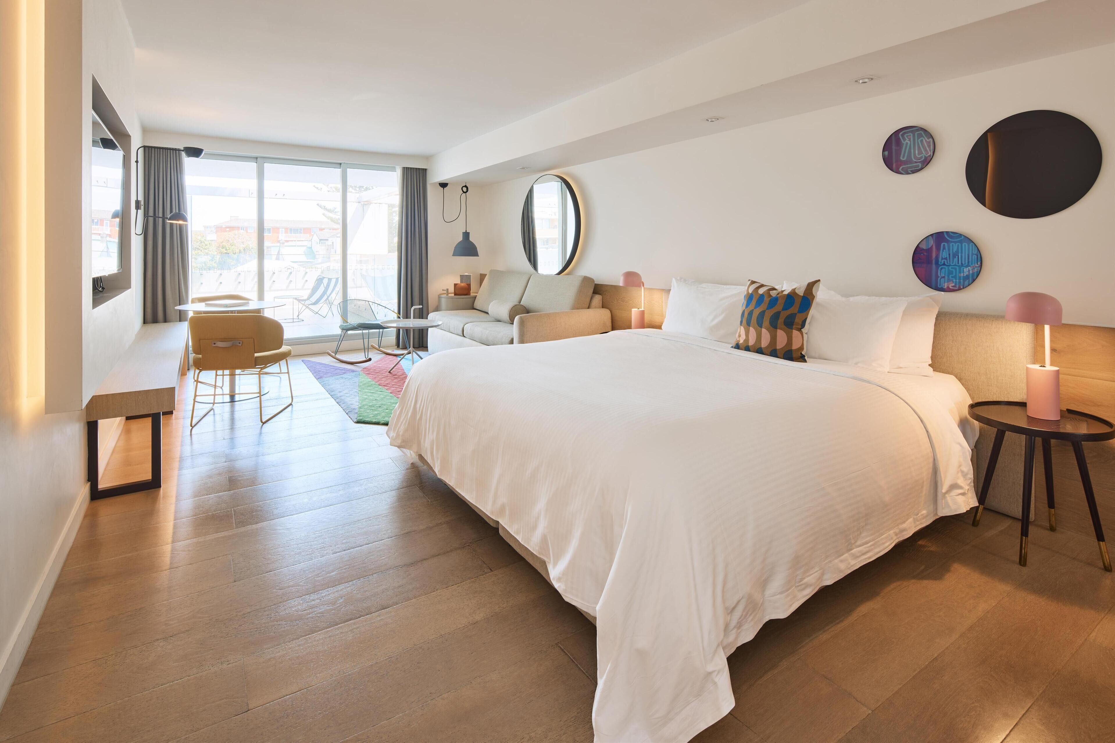 A modern hotel room with a large bed, a desk, and a chair. There is a large window providing a view of the city.