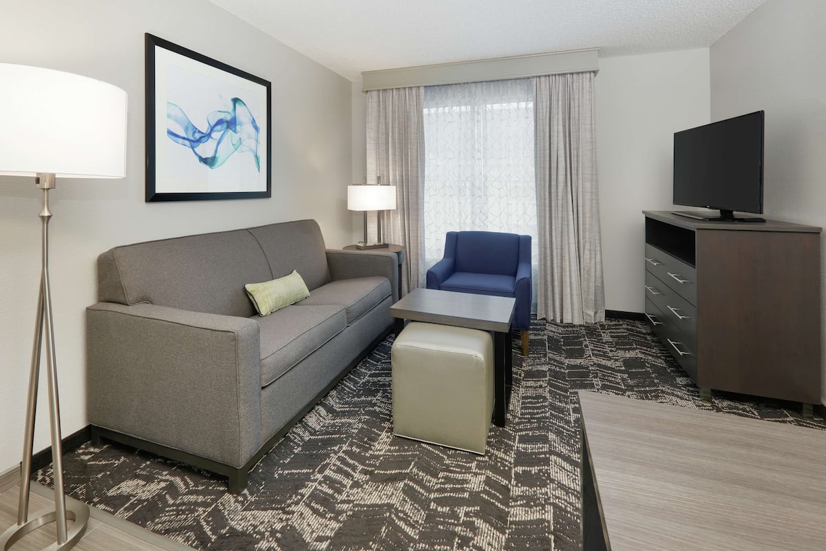 Una habitación de hotel con un sofá gris, un sillón azul, un pequeño otoman, un televisor sobre un mueble de madera y una alfombra estampada.