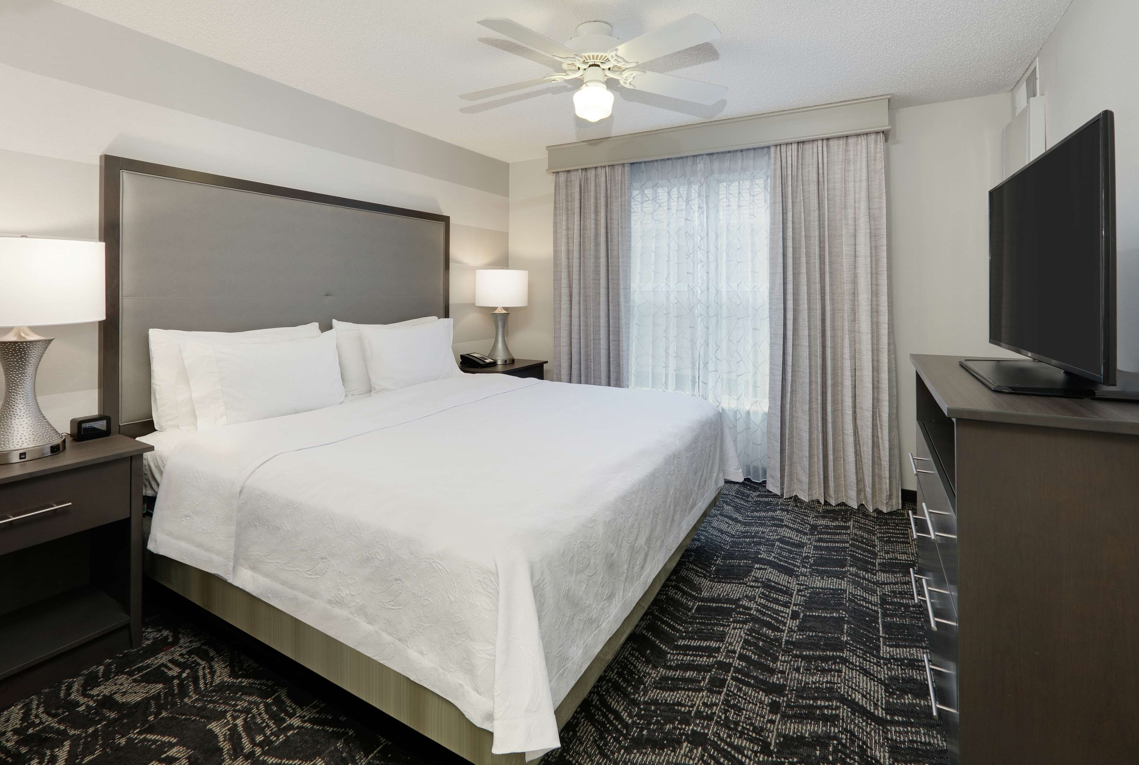 A hotel room with a large bed, a ceiling fan, a flat-screen TV, and a patterned rug.