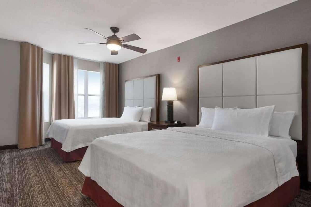 A hotel room with two beds, a ceiling fan, a window with curtains, and a nightstand with a lamp.