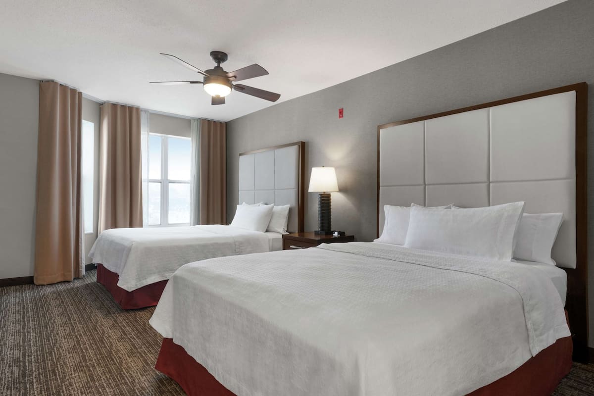 A hotel room with two beds, a ceiling fan, a nightstand with a lamp, and a window with curtains.