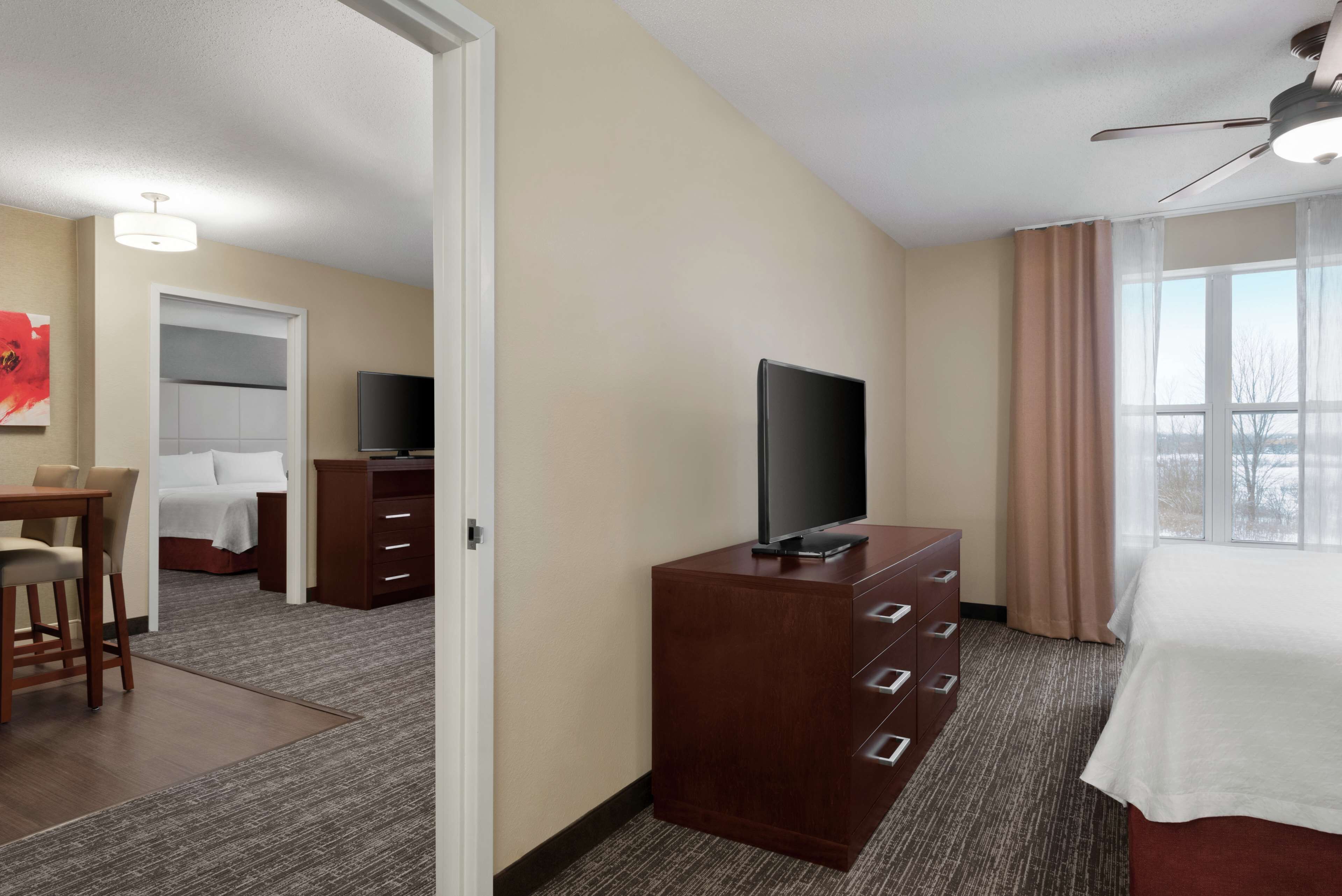 A hotel room with a bed, a TV on a wooden dresser, a desk with a chair, and a window with curtains.