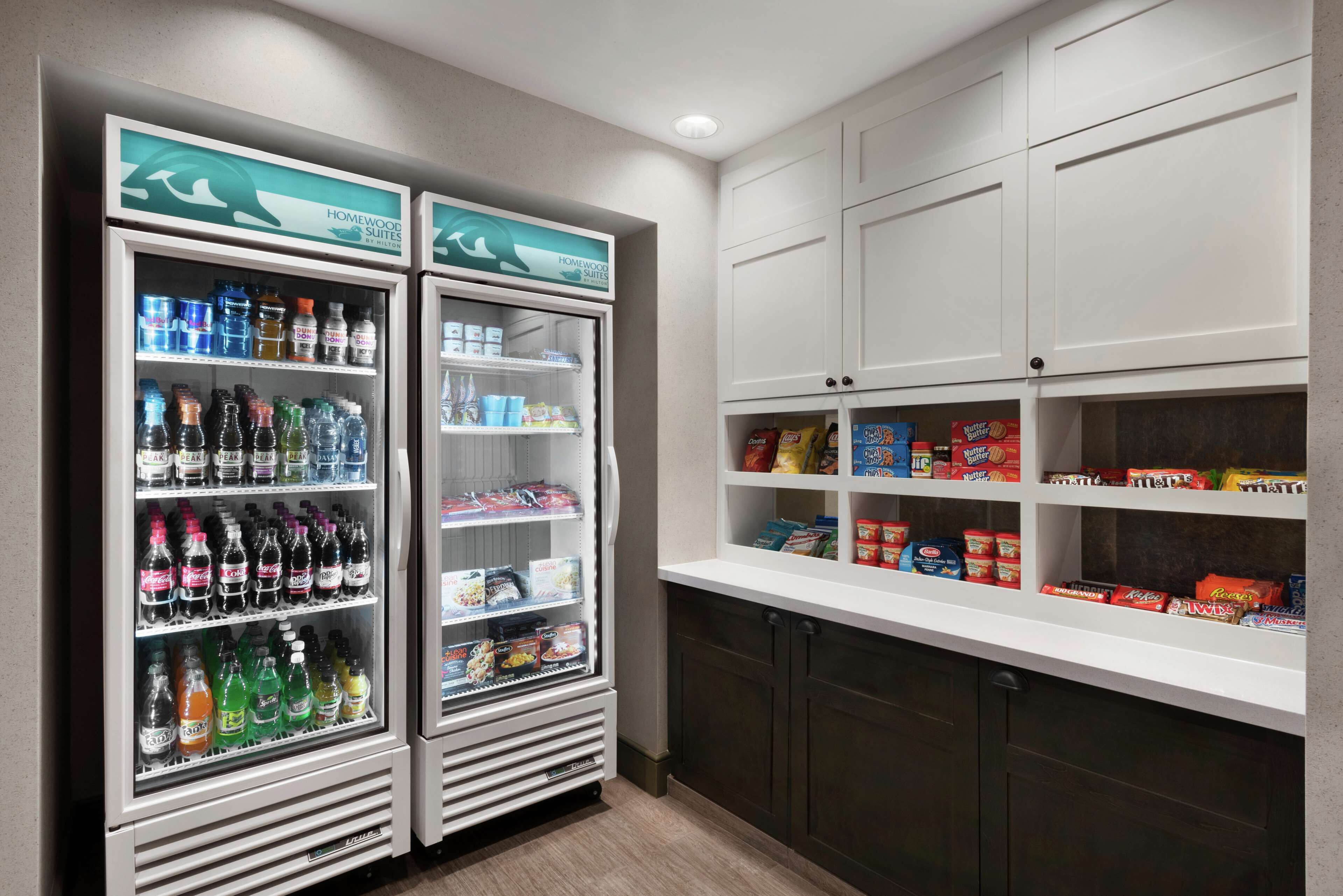 A kitchenette with two beverage dispensers stocked with various drinks, a white countertop with snacks and condiments, and white cabinetry.