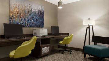 A hotel room with a desk, two yellow chairs, a teal chair, a printer, a computer, and a large abstract painting on the wall.