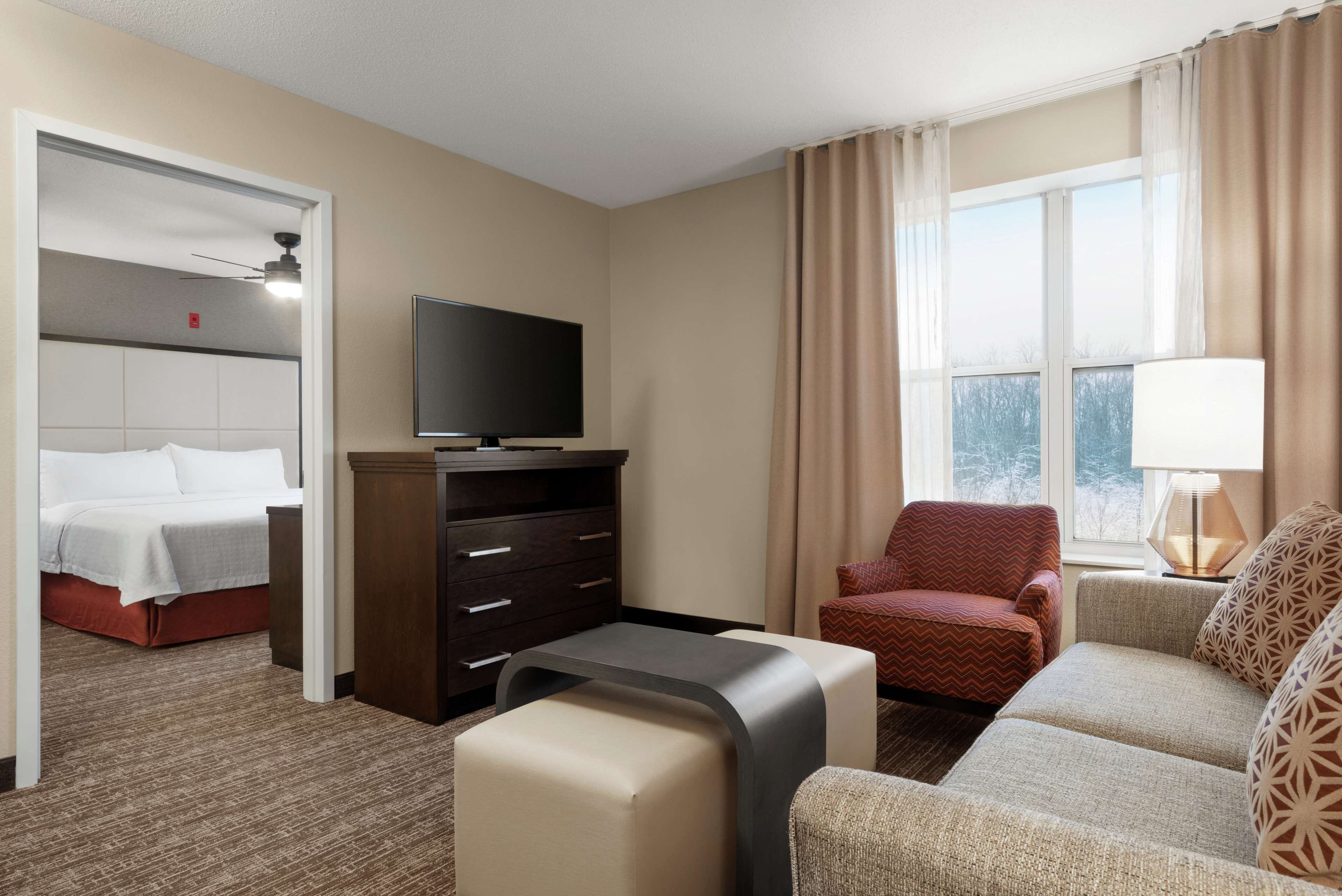 A hotel room with a bed, a TV, a desk, a chair, a sofa, and a window with curtains.