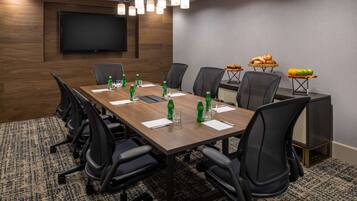 Un salón de conferencias con una larga mesa, sillas, un televisor de pantalla plana y una mesa pequeña con refrigerios.