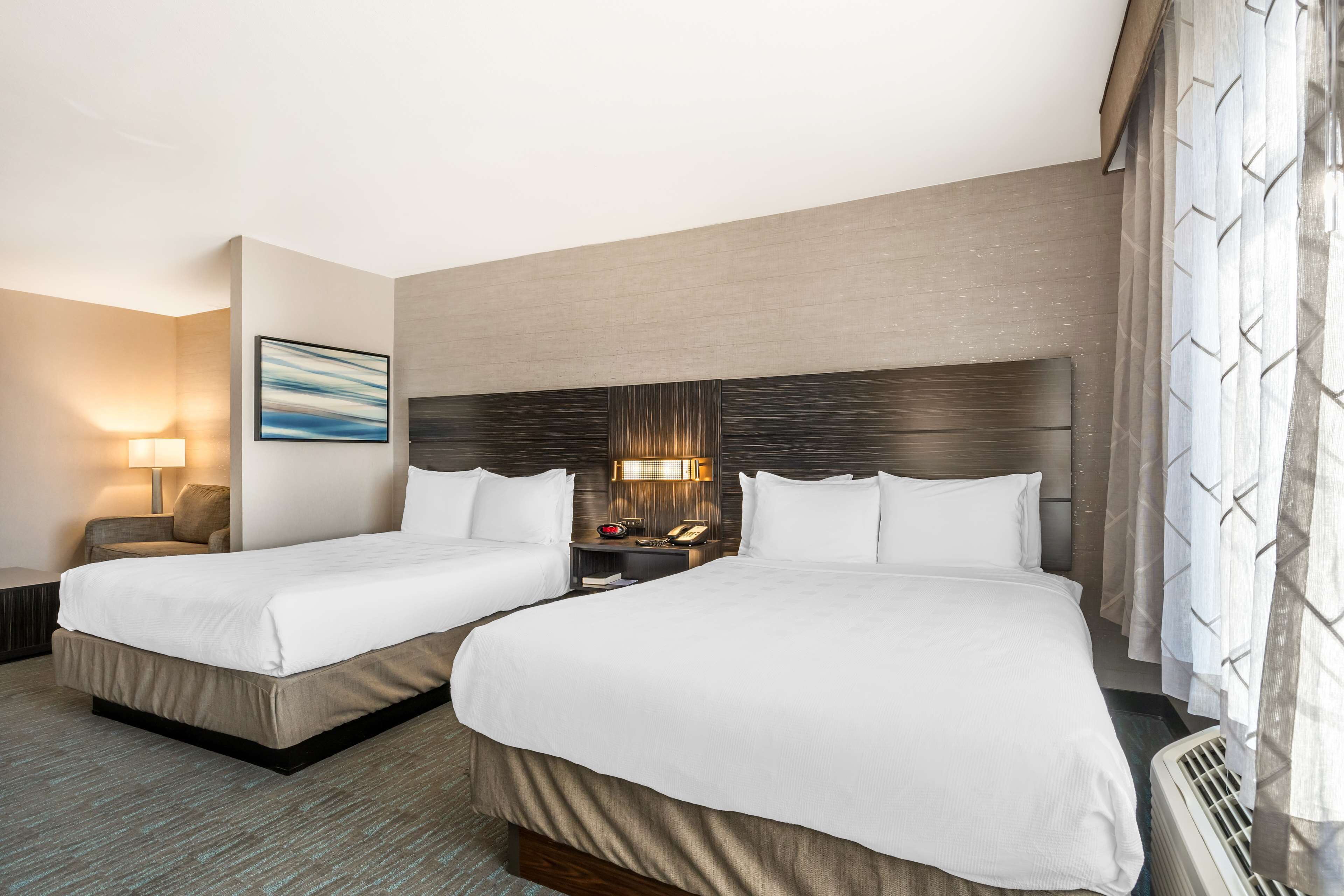 A hotel room with two beds, a nightstand with a lamp, a painting on the wall, and a window with curtains.