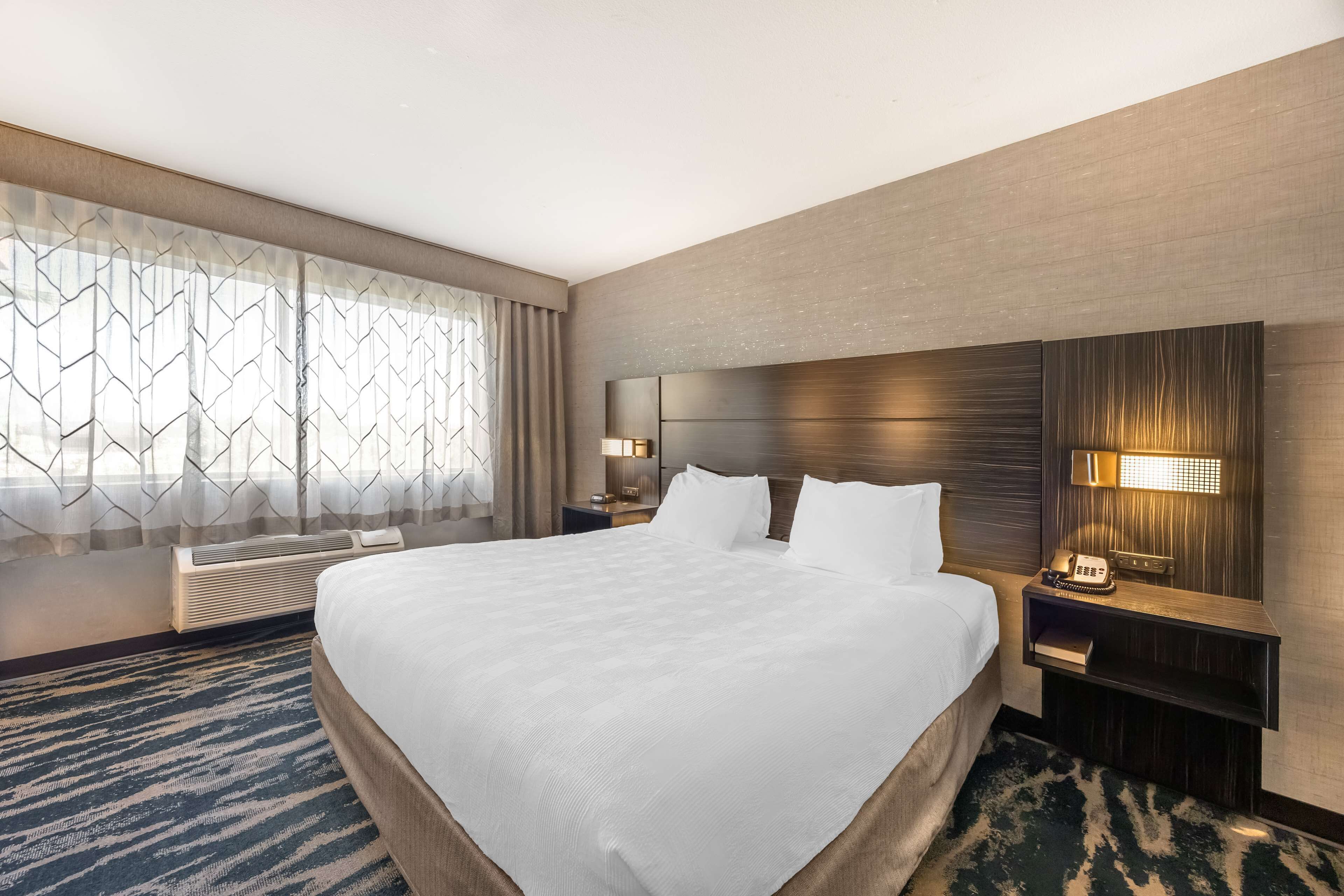 A hotel room with a large bed, a nightstand with a lamp, a wall-mounted phone, and a window with a geometric curtain.