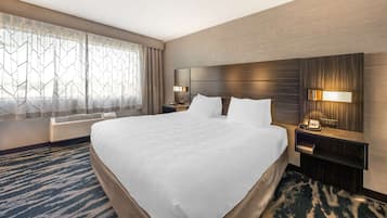 A hotel room with a large bed, a nightstand with a lamp, a wall-mounted phone, and a window with a geometric curtain.