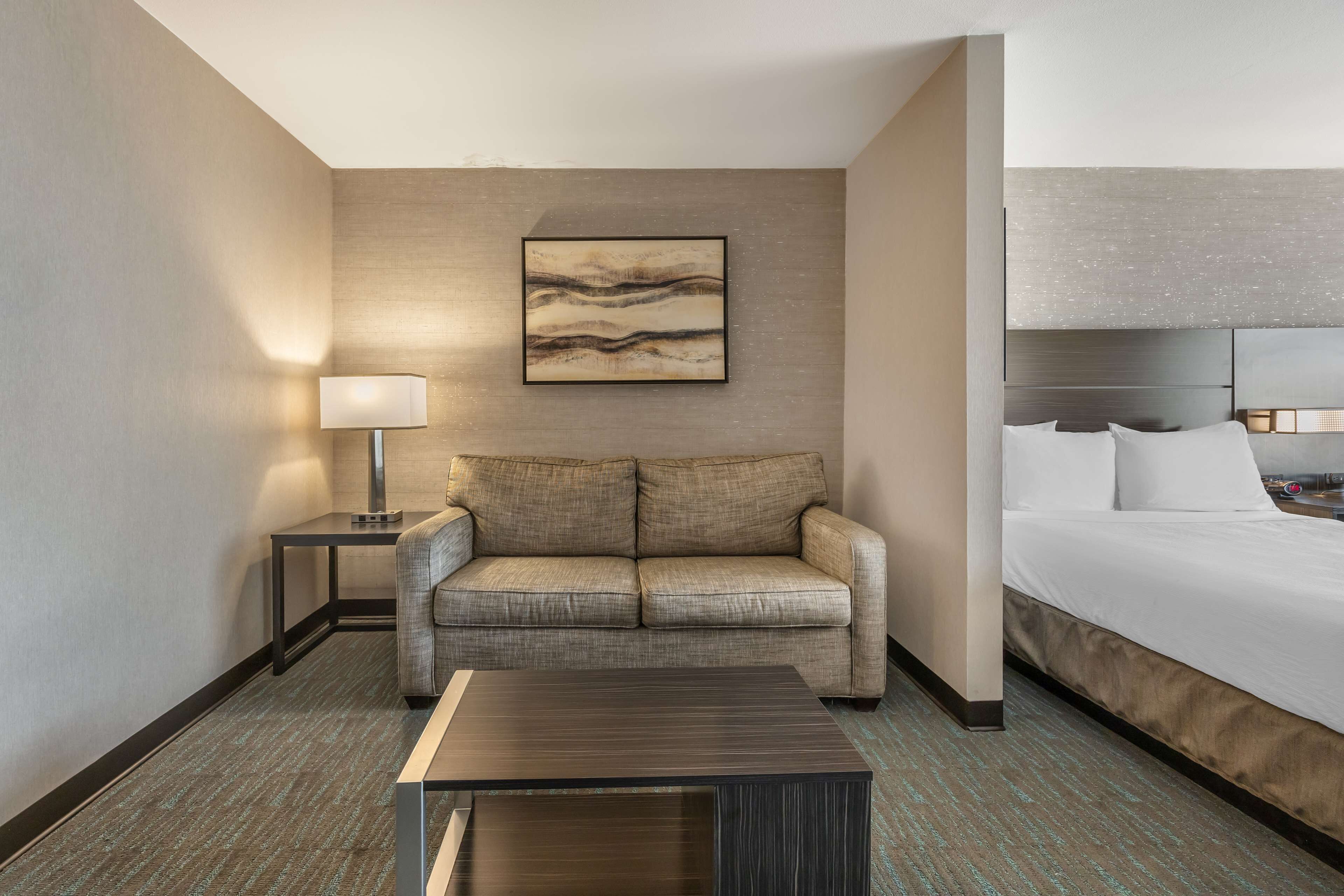 A hotel room with a sofa, a bed, a bedside table, a lamp, and a wall-mounted artwork.