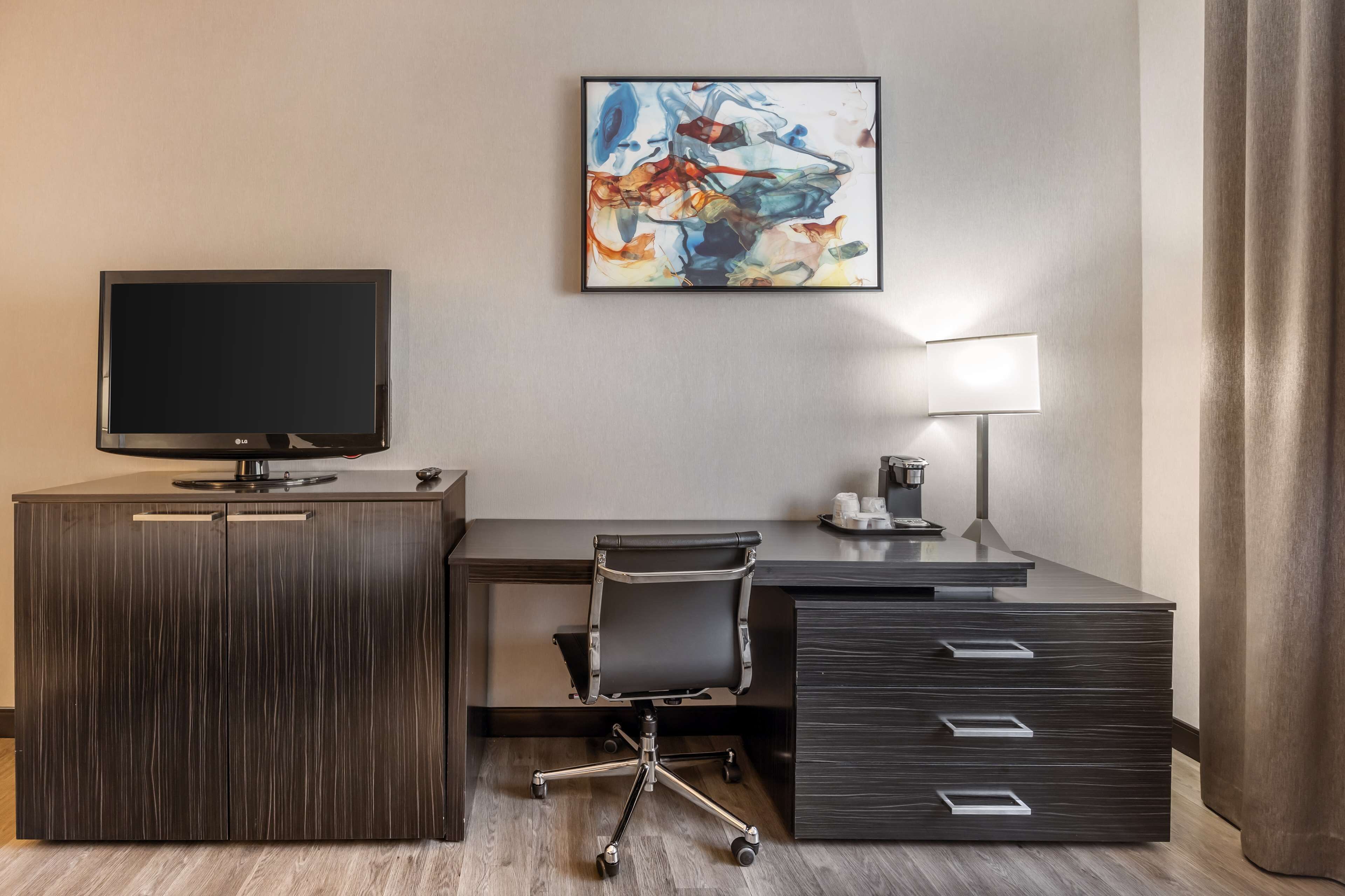 A hotel room with a dark wood desk, a flat-screen TV, a modern chair, a desk lamp, and a large abstract painting on the wall.