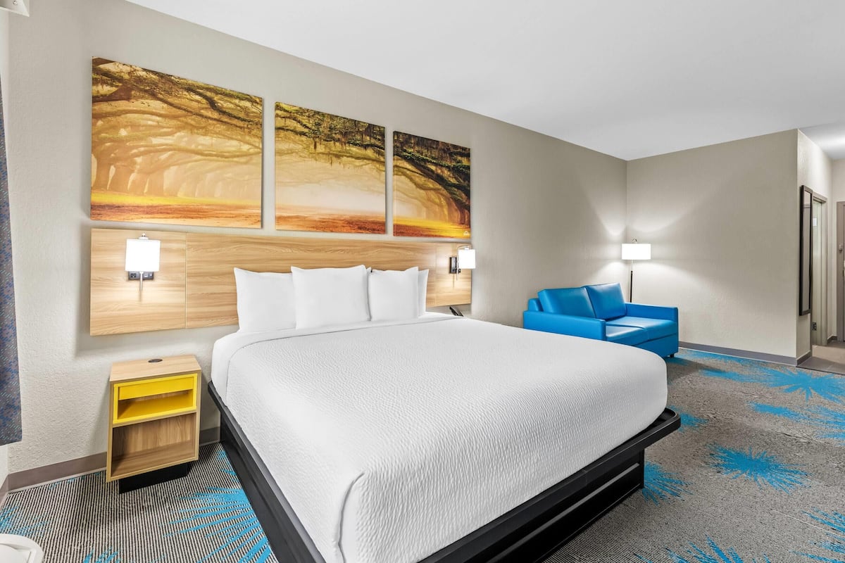 A hotel room with a large bed, a nightstand, a blue chair, and a wall-mounted artwork.