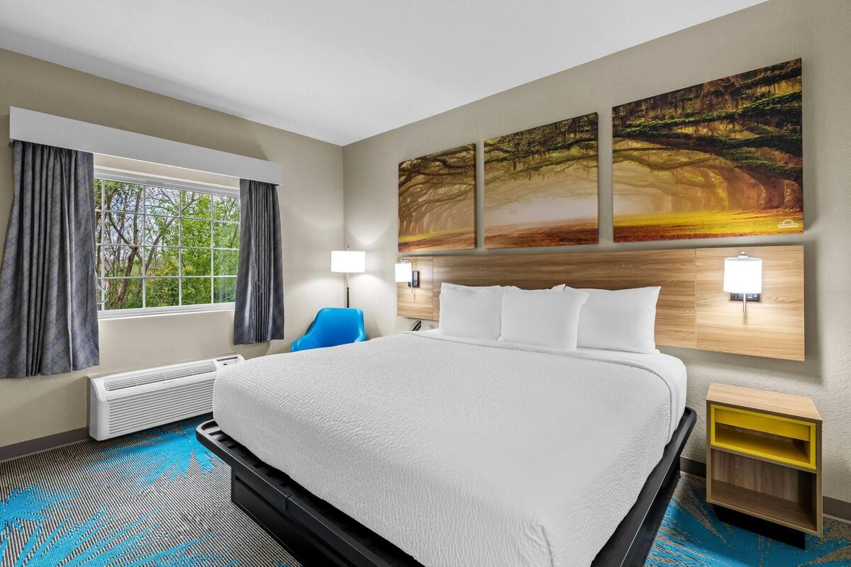 A hotel room with a large bed, a window with curtains, a blue chair, a wall-mounted lamp, and a painting.