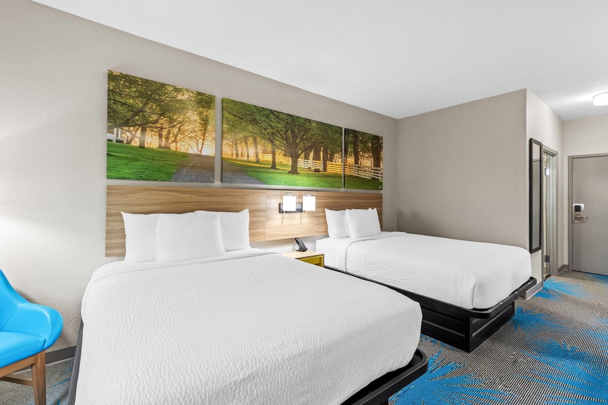 A hotel room with two beds, a large wall painting, a desk with a lamp, and a blue chair.