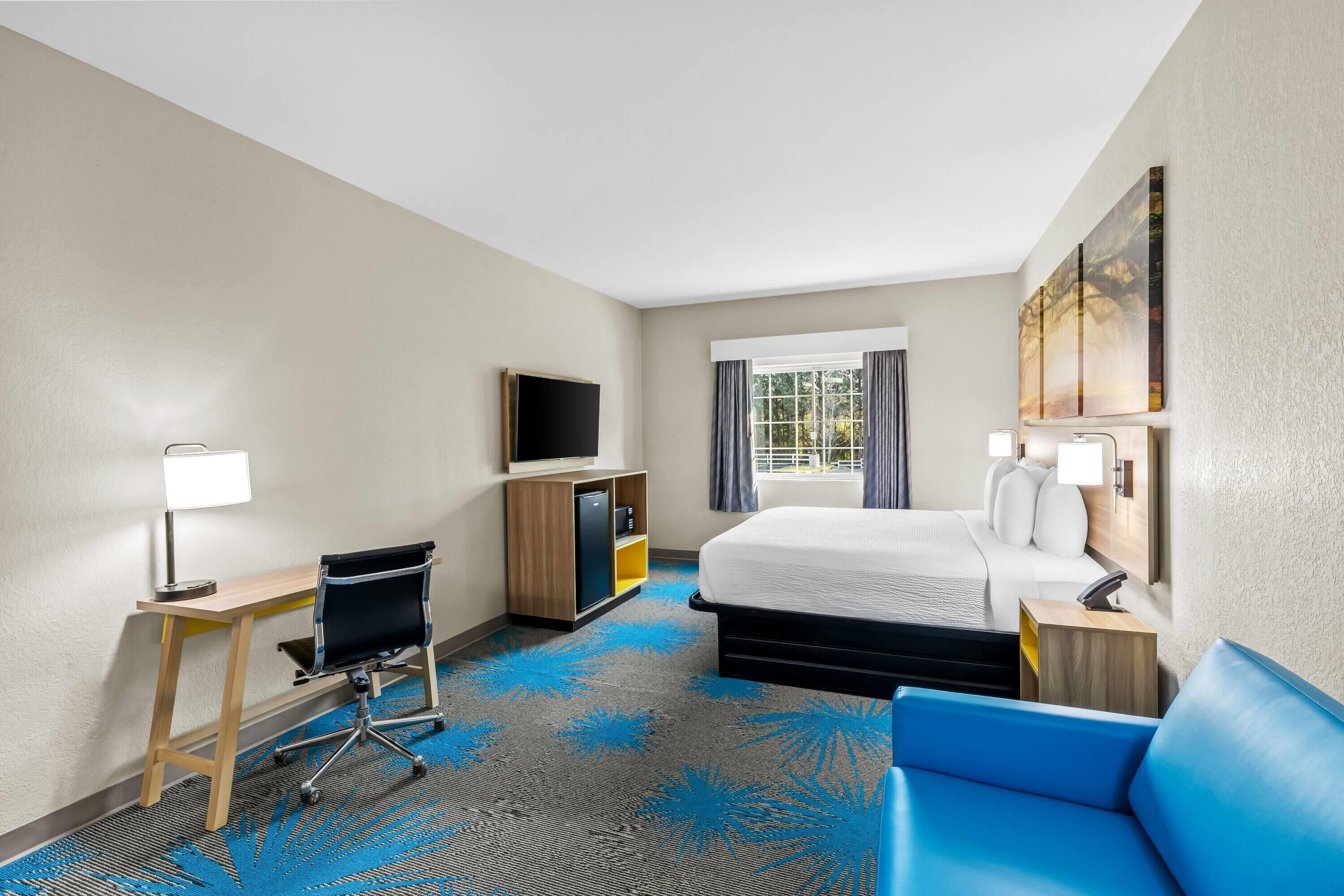Habitación de hotel con cama, escritorio, silla, televisor y ventana con cortinas.