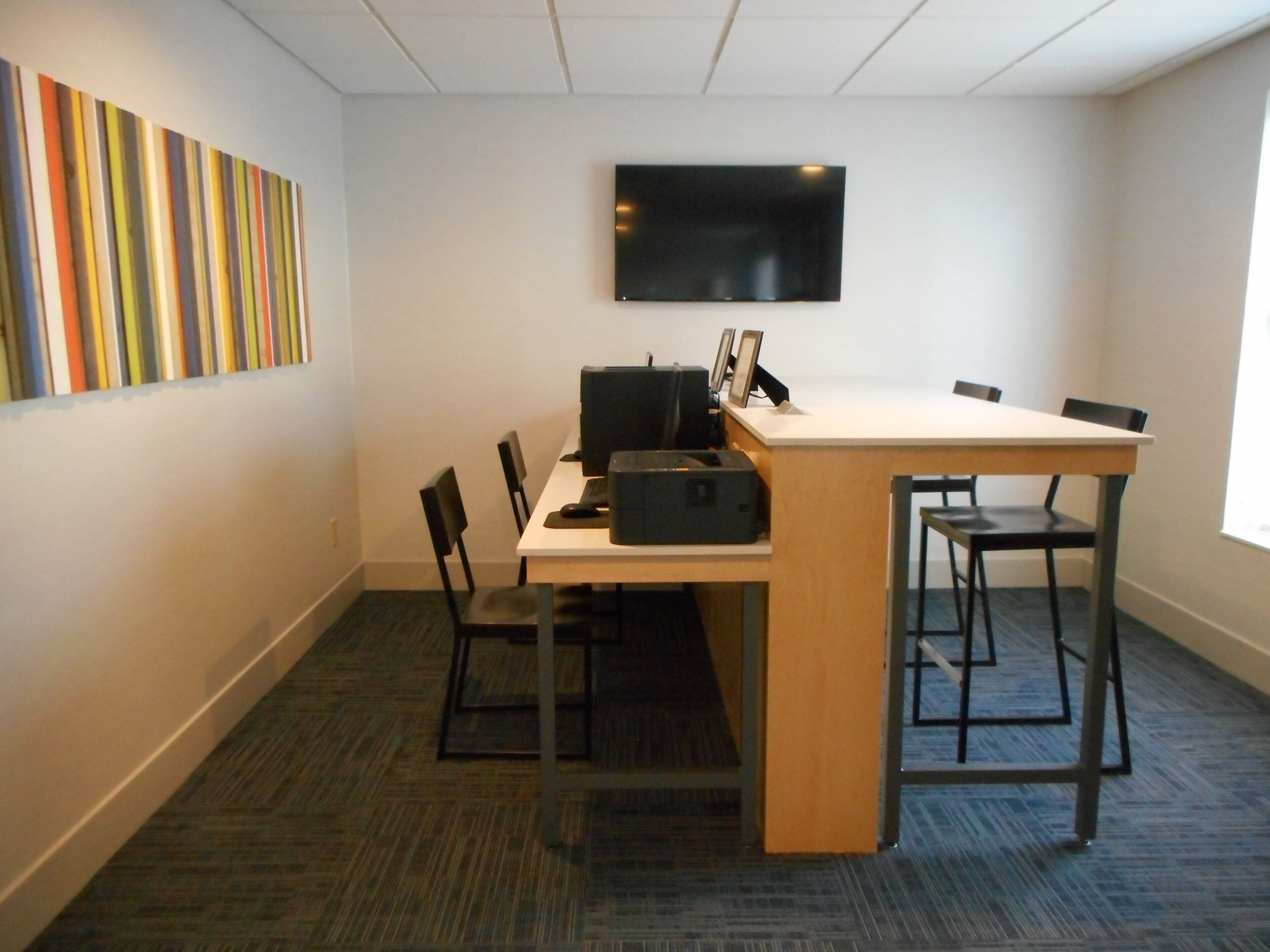 Une salle de conférence avec une longue table, quatre chaises, un téléviseur à écran plat fixé au mur et une décoration murale colorée à rayures.