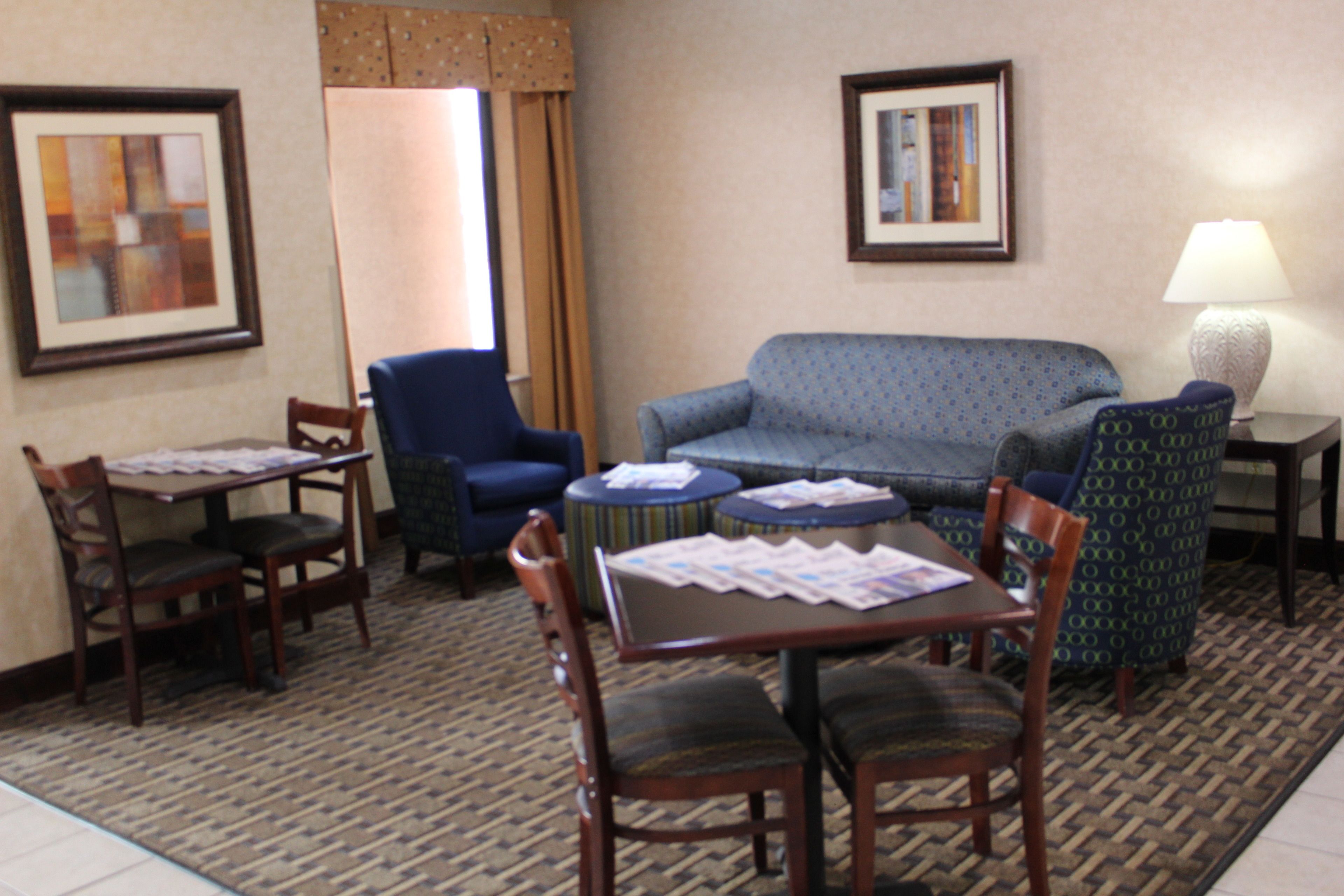 Habitación de hotel con un sofá azul, dos sillones, una mesa de comedor de madera con sillas, una mesita redonda con una lámpara y dos cuadros en las paredes.