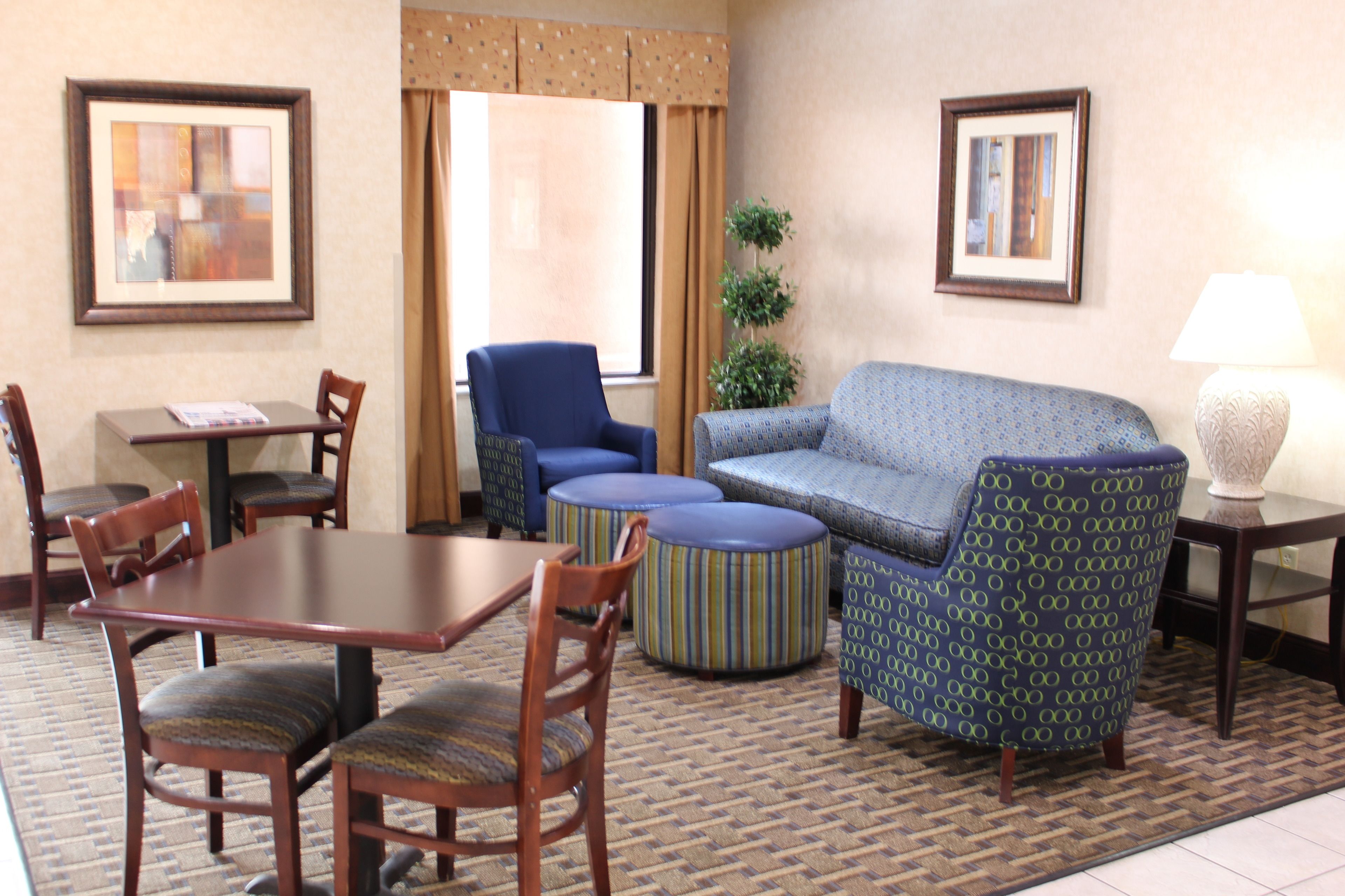 Habitación de hotel con una mesa de comedor, sillas, sofá, otomanas, lámpara y cuadros en las paredes.