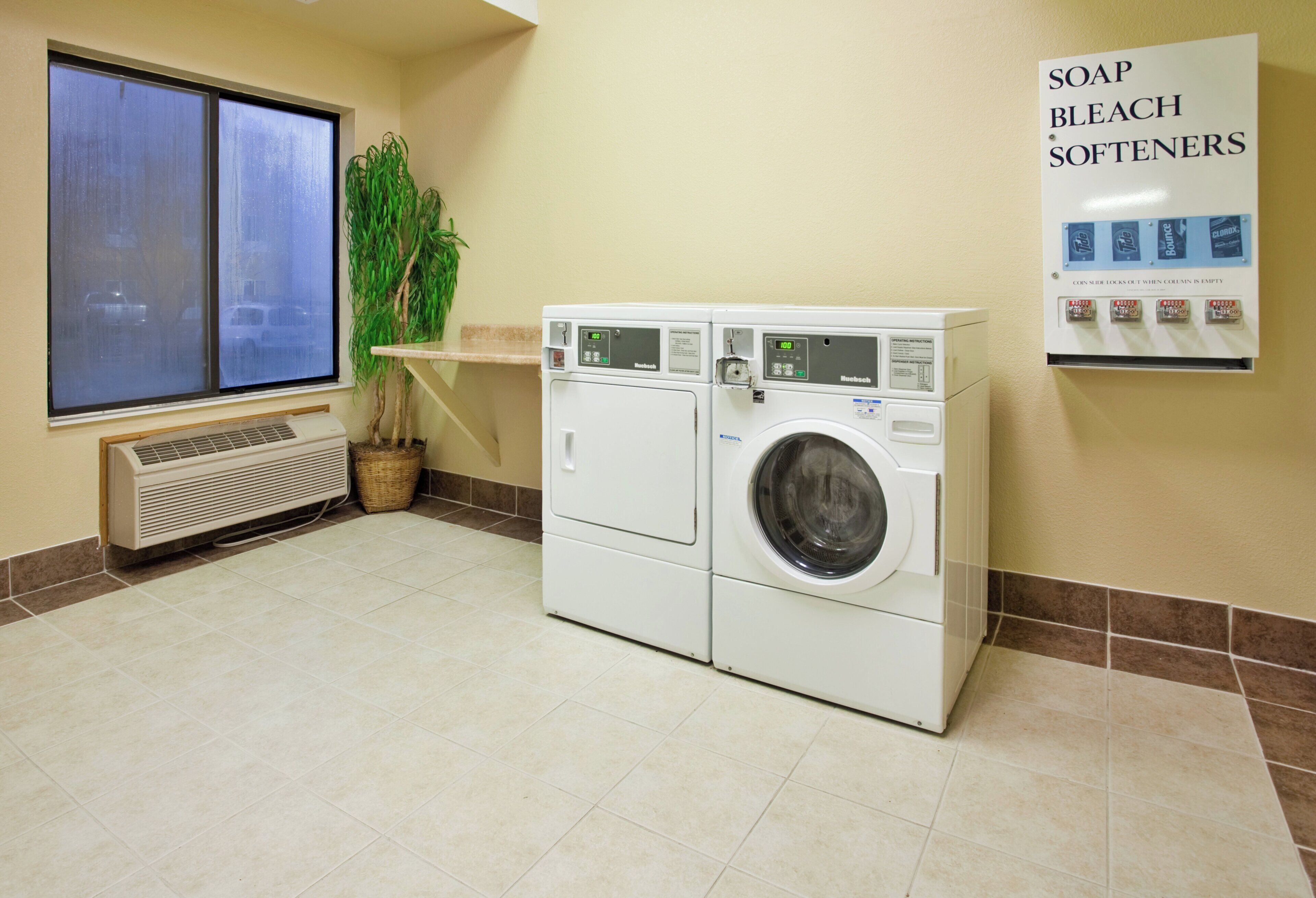 Un lavadero con lavadora y secadora apiladas, una ventana con persianas, una planta en maceta y un cartel que enumera los detergentes disponibles.