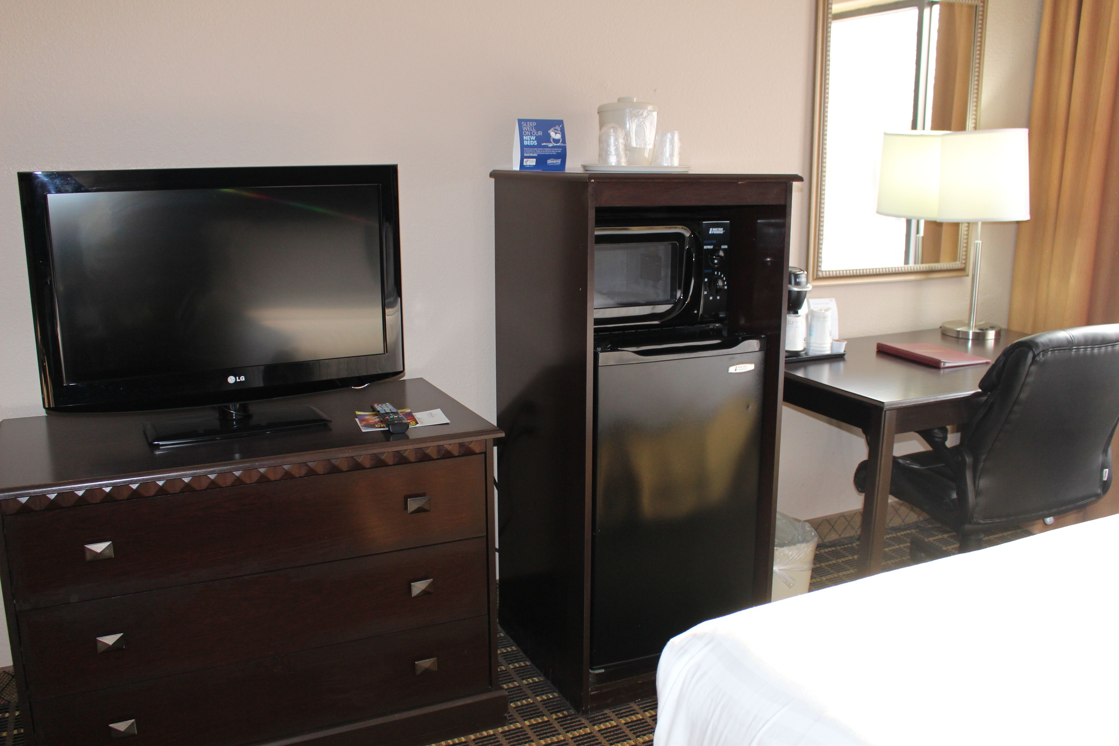 A hotel room with a flat-screen TV, a microwave, a refrigerator, a desk with a lamp, and a chair.