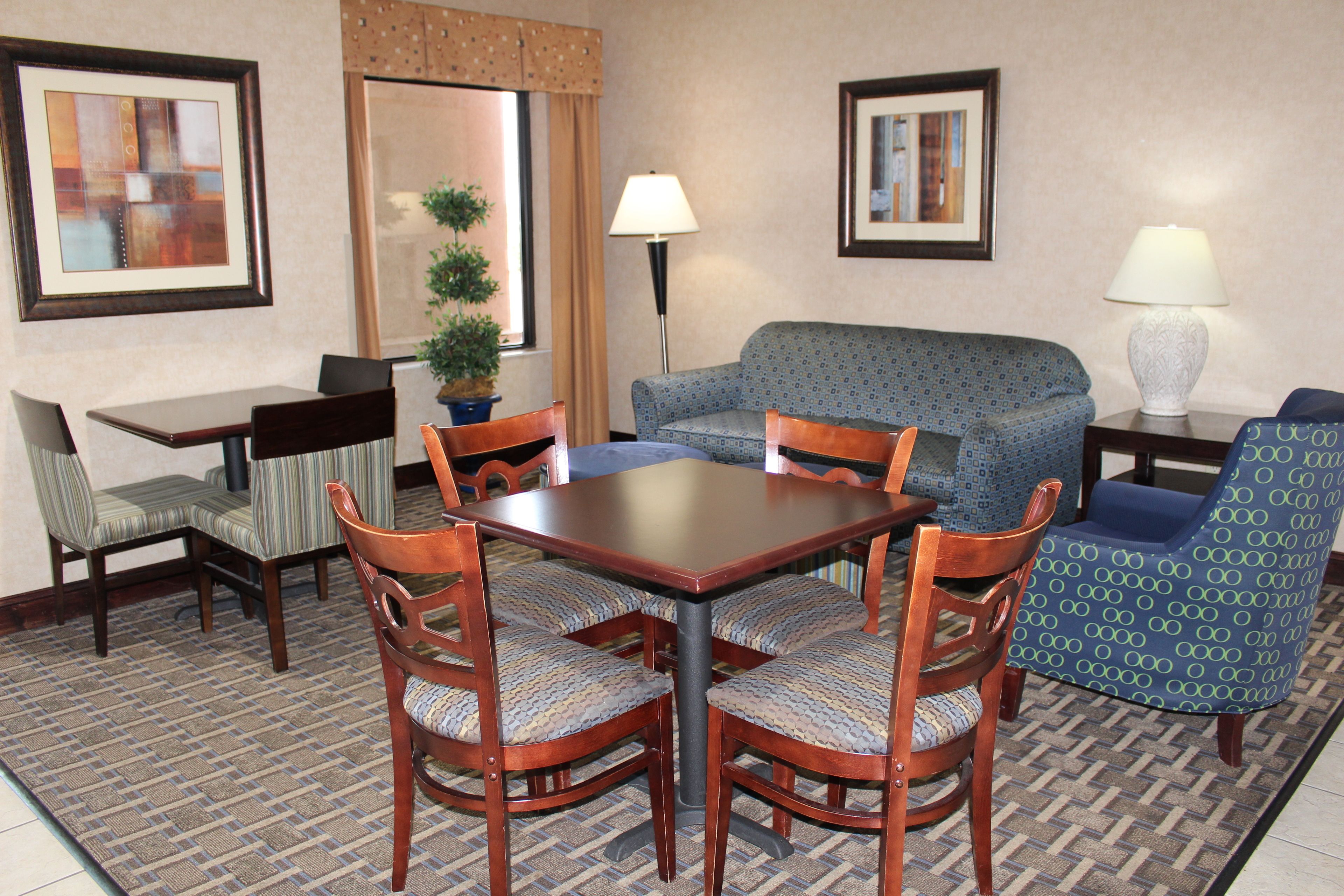 Habitación de hotel con una mesa de comedor, cuatro sillas, un sofá, una lámpara y dos cuadros en las paredes.