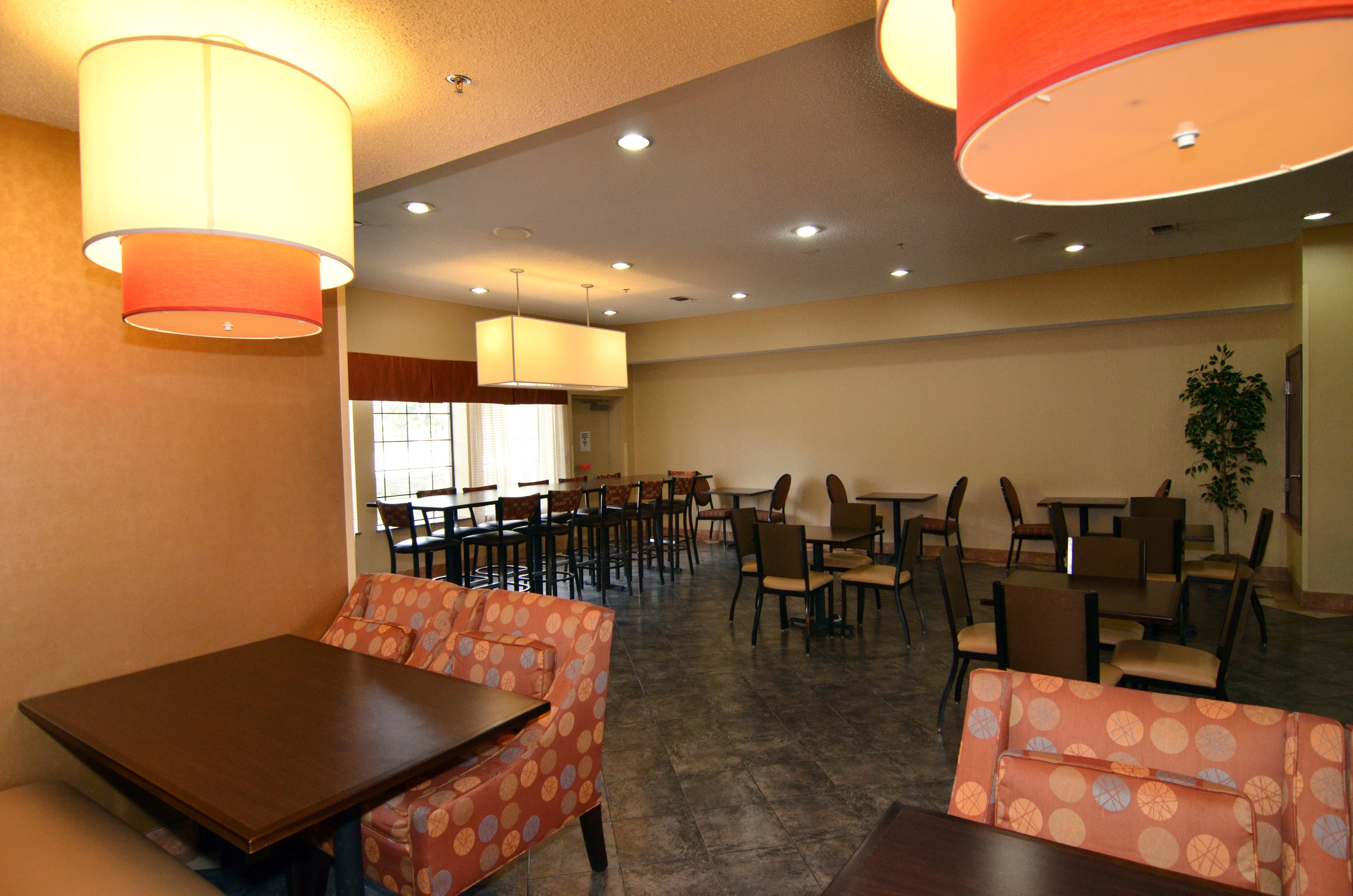 Un comedor espacioso con varias mesas y sillas, que presenta sofás color naranja con estampado y un gran ventanal que proporciona luz natural.