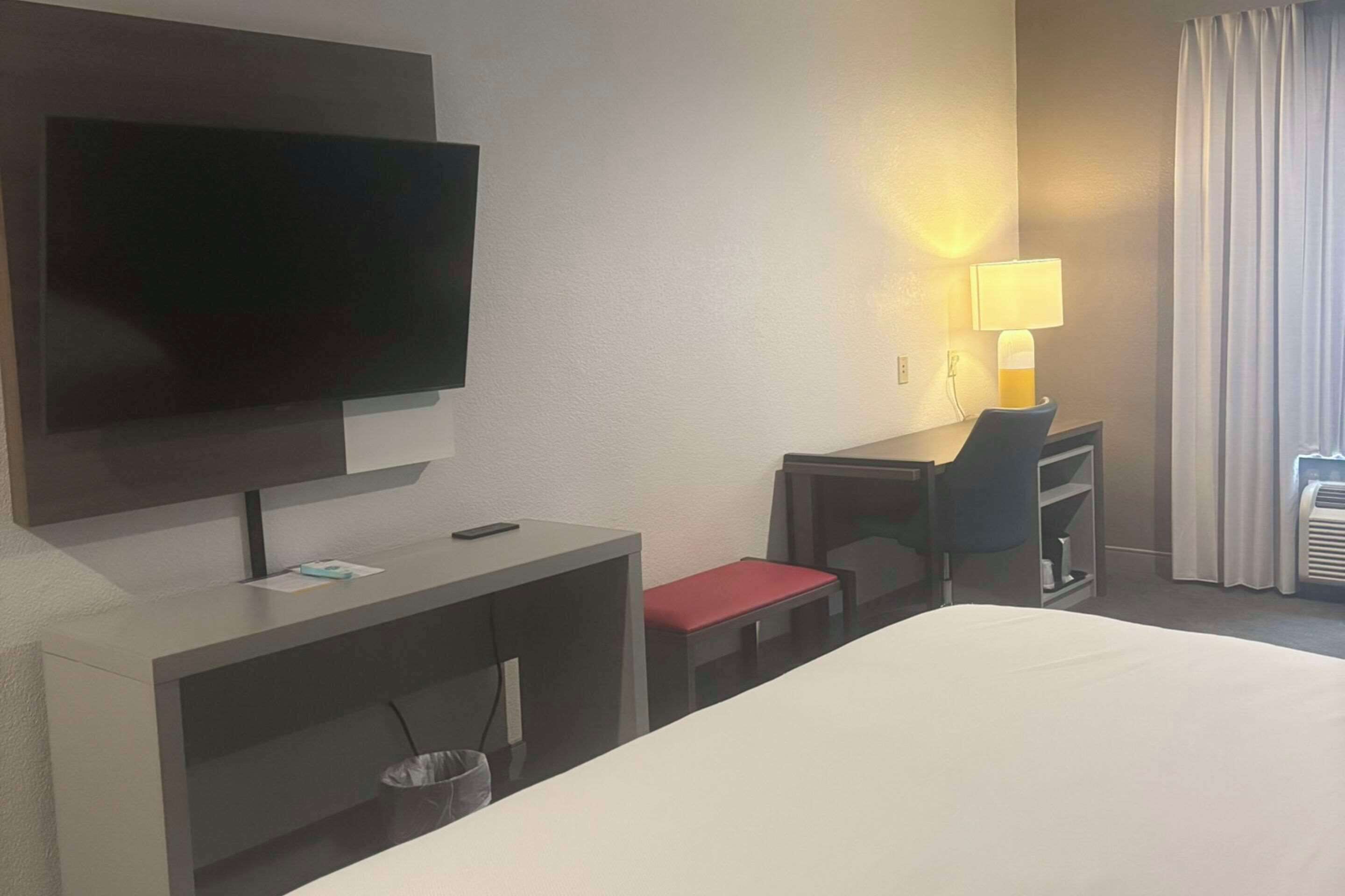 A hotel room with a bed, a desk, a chair, a television, a lamp, and a window with curtains.