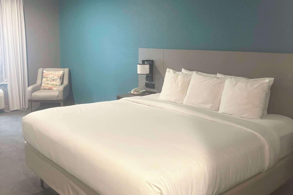 A neatly made bed with white linens, a chair with a floral pillow, a bedside lamp, and a phone on a nightstand.
