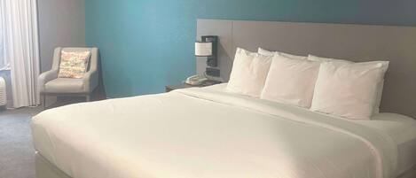 A neatly made bed with white linens, a chair with a floral pillow, a bedside lamp, and a phone on a nightstand.