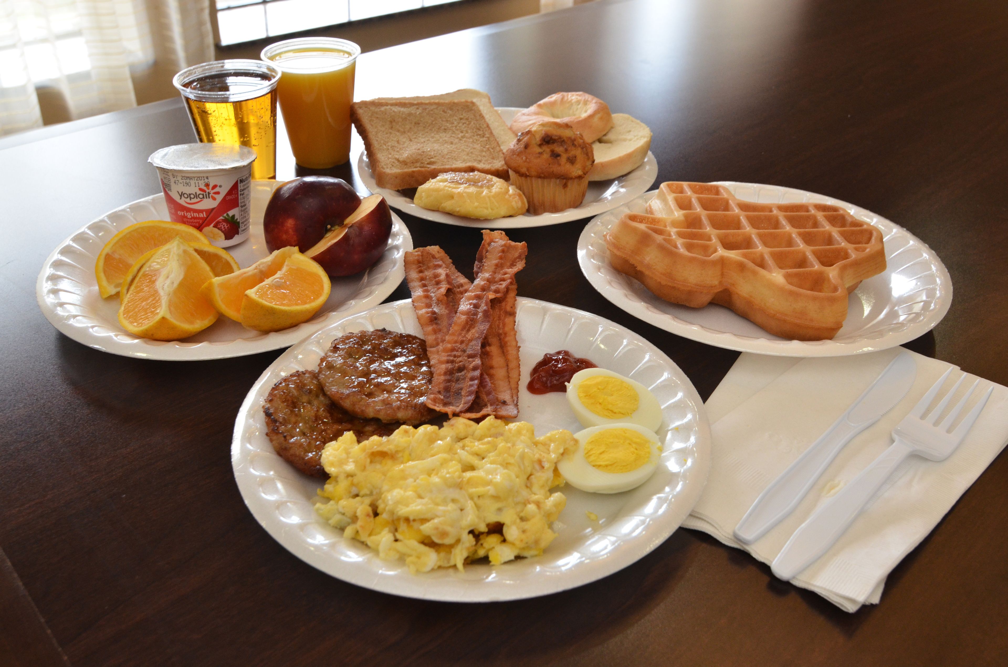 Une table de petit-déjeuner avec des assiettes remplies d’œufs, de bacon, de pain grillé, de gaufres et des fruits.