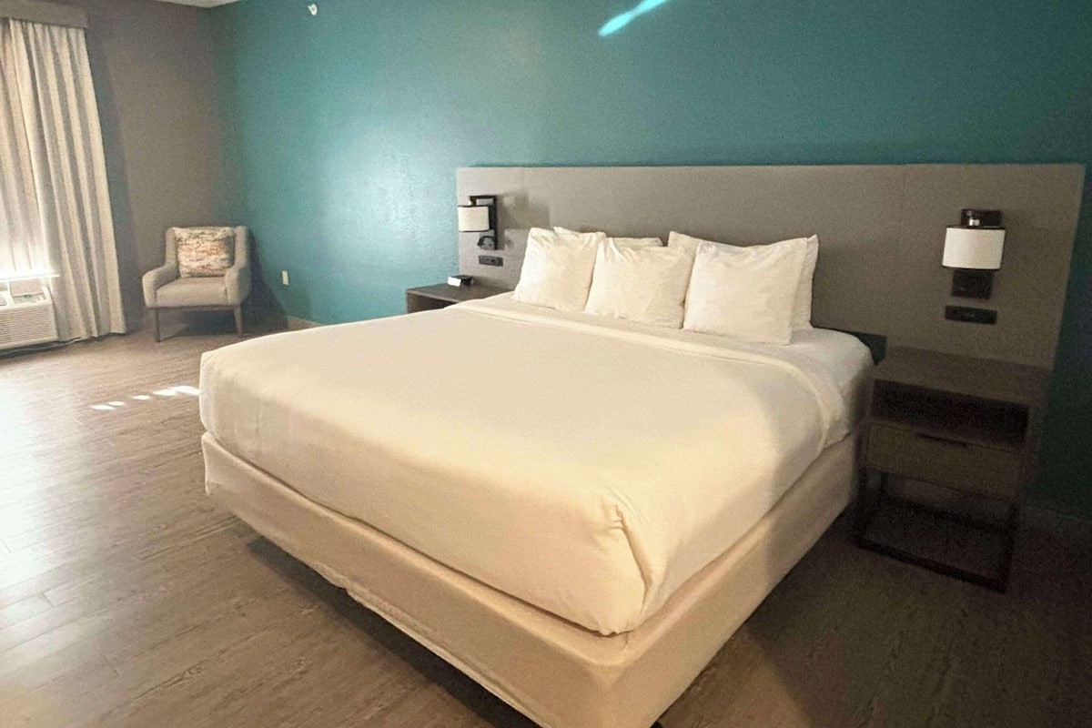 A large bed with a headboard, two bedside tables, and a chair in a room with teal walls and wooden flooring.