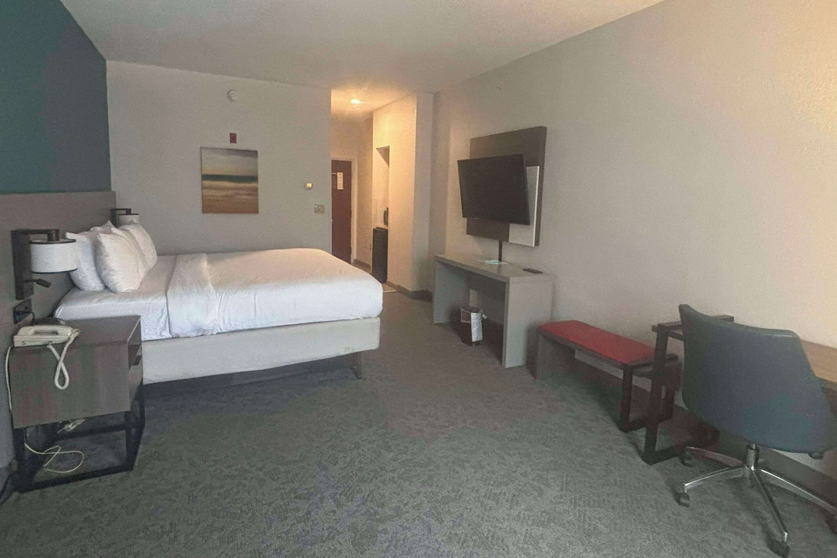 A hotel room with a bed, a desk with a telephone, a chair, a television on a stand, and a painting on the wall.
